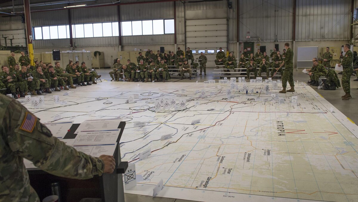 Des militaires participent à un exercice de stratégie. Une énorme carte est étalée sur le sol avec des pions posés dessus. Un des militaires pointe quelque chose sur la carte alors que les autres regardent.
