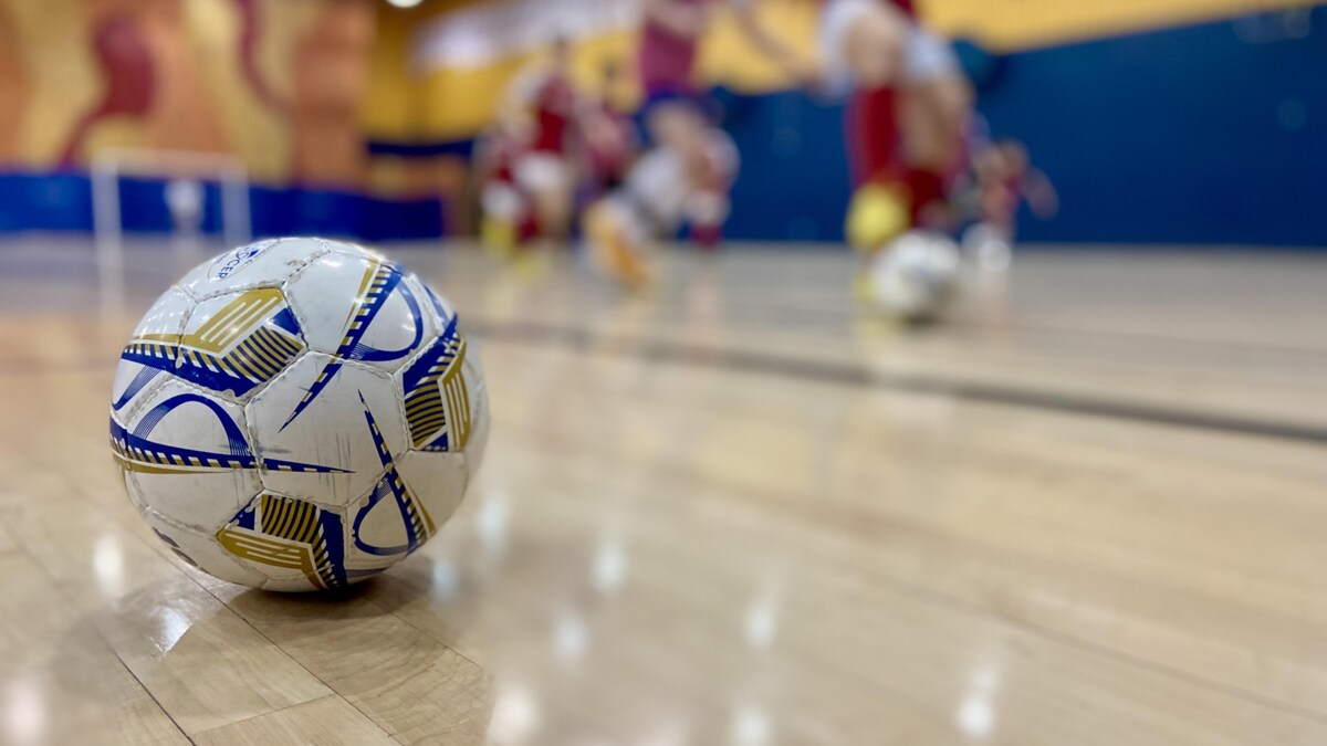 Gros plan sur un ballon de soccer dans un gymnase, pendant que des jeunes jouent derrière. 