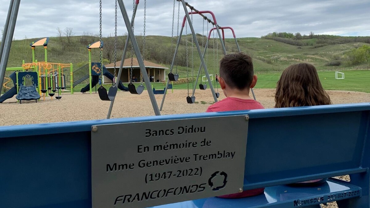 Deux enfants sont assis sur un banc en métal devant un terrain de jeu au printemps au Manitoba. Sur le banc, il est écrit «Bancs Didou. En mémoire de Mme Geneviève Tremblay (1947-2022) Francofonds».