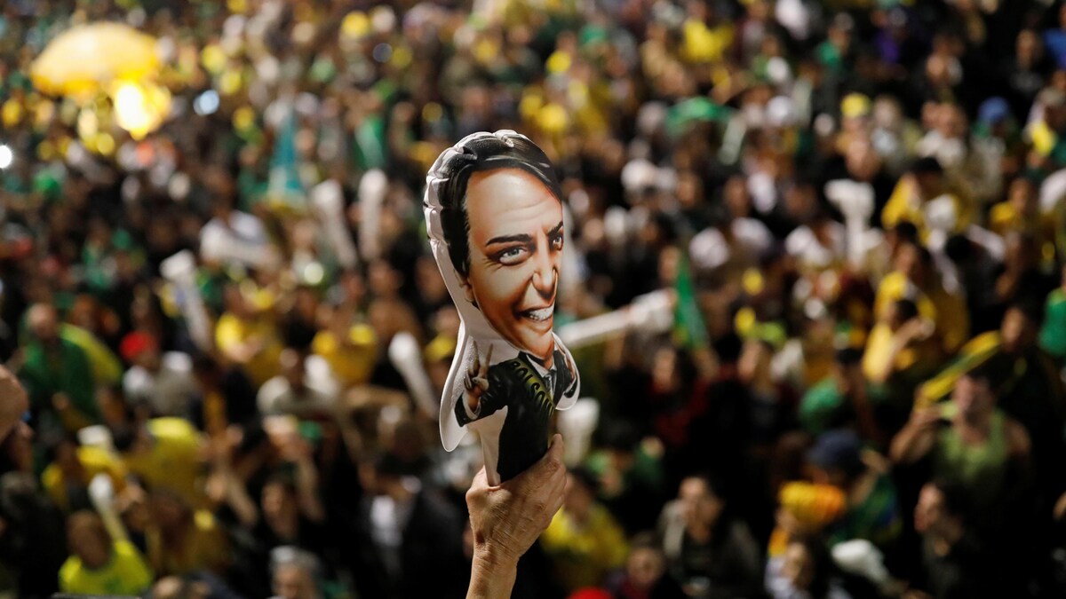 Une foule immense est réunie en arrière-plan. Le focus de la photo est fait sur une petite affiche représentant Bolsonaro.