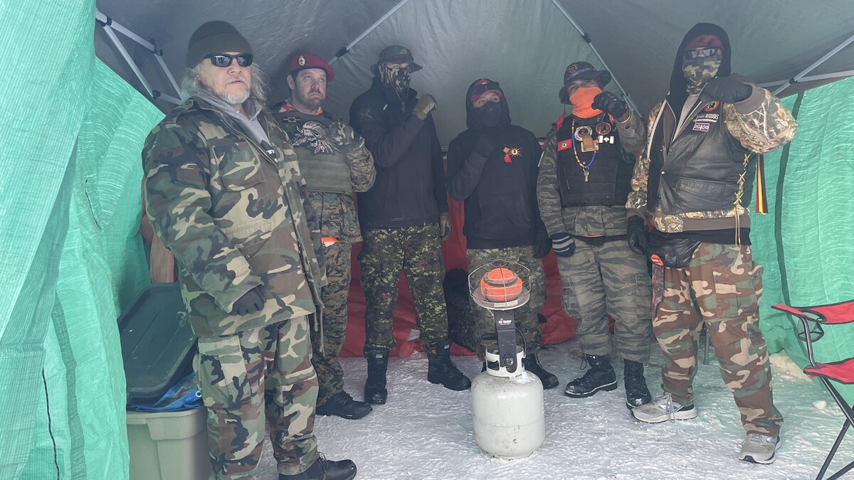 Six hommes vetues en uniforme de camouflage sous un campement. 
