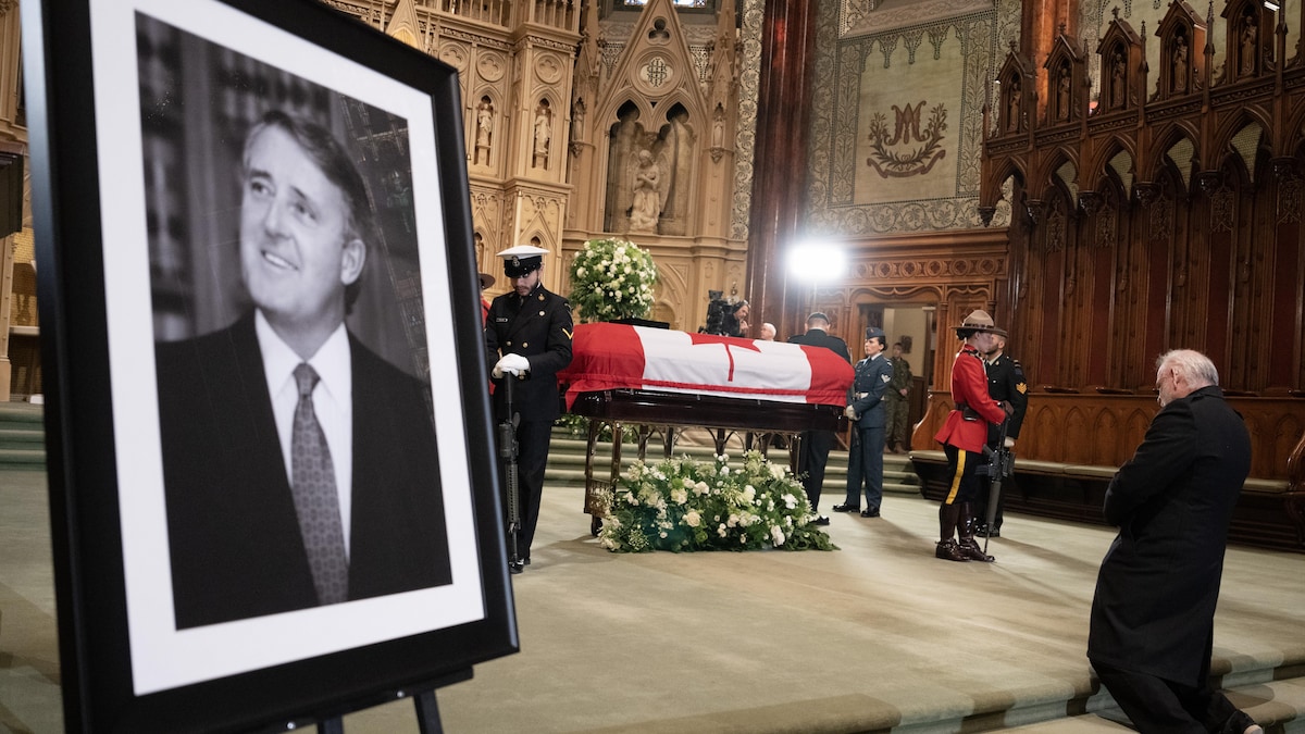 Une photo de Brian Mulroney est exposée dans la basilique, où un homme est agenouillé devant le cercueil recouvert du drapeau du Canada et entouré de policiers de la GRC et de membres de l'armée qui le veille.