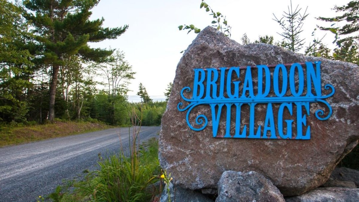 Une enseigne bleue « Brigadoon Village » décore une grosse roche près d'un chemin rural menant au camp.