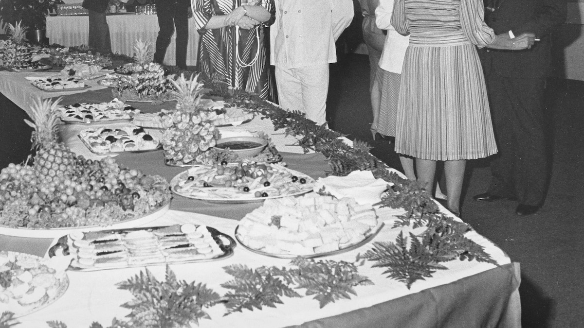 Un chef cuisinier pose en compagnie d'une invitée près d'un buffet avec plusieurs grands plateaux de nourriture décorés d'ananas entier.
