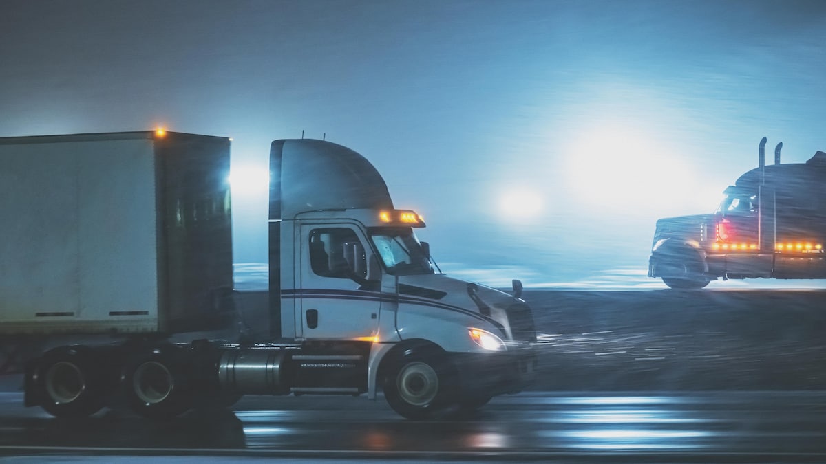 Deux camions semi-remorques circulent en direction opposée sur deux voies d'une autoroute, la nuit, dans la pluie et le grésil.