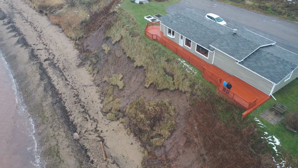 Deux personnes sur la galerie d'une maison examinent les dégâts de l'érosion en contrebas. Des portions du terrain sont tombées sur la plage.