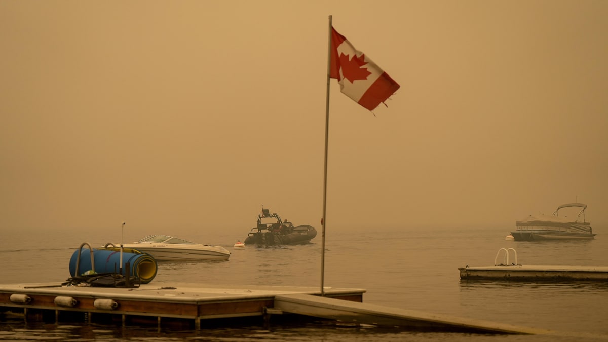 L'épaisse fumée qui se dégage du feux de Lower East Adams Lake, en Colombie-Britannique, enveloppe un drapeau canadien et des bateaux sur le lac.