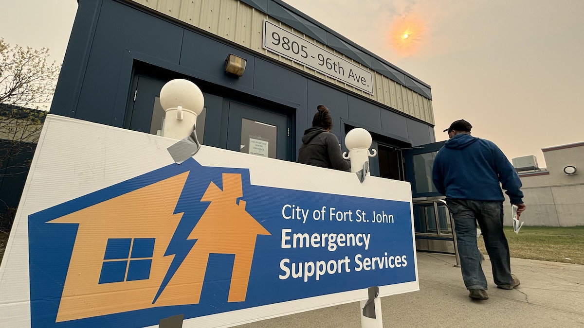 Deux personnes entrent dans le centre d'évacuation. Une affiche de la ville de Fort St John qui explique qu'il s'agit de services d'urgence a été installée devant l'entrée. 