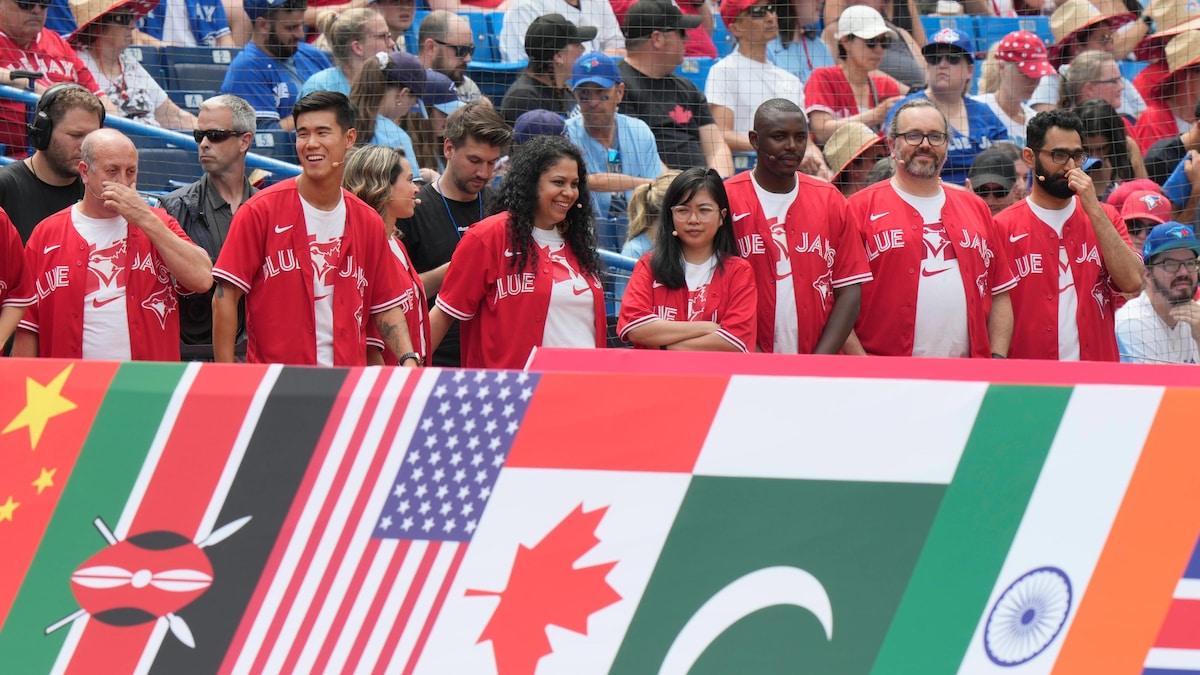 Sur un terrain de baseball, une rangée de personnes portent des chemises rouges des Blue Jays et sont en rang devant de grandes répliques des drapeaux de la Chine, du Kenya, des États-Unis, du Canada, du Pakistan, de l'Inde et de la Grande-Bretagne.