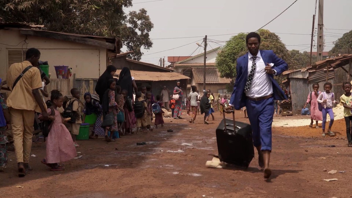 Un homme africain vêtu d'un habit marche sur une rue de terre avec une valise.
