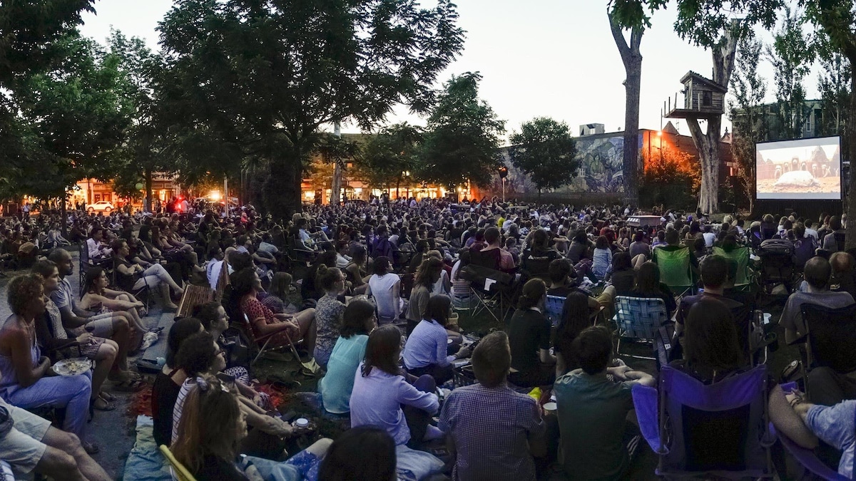 Une foule de personnes assises sur des chaises pliantes regardent un écran de cinéma installé dans un parc.