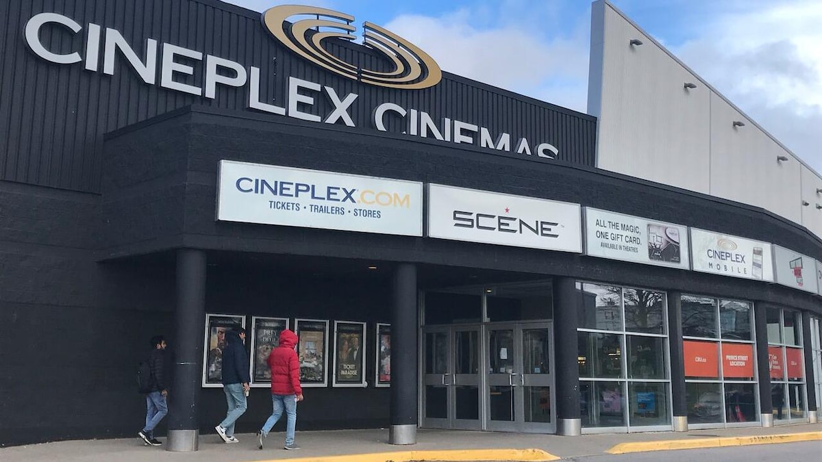 La structure extérieure d'une salle de cinéma Cineplex à Sydney, en Nouvelle-Écosse. Trois personnes de dos en manteau marchent vers l’entrée principale. 
