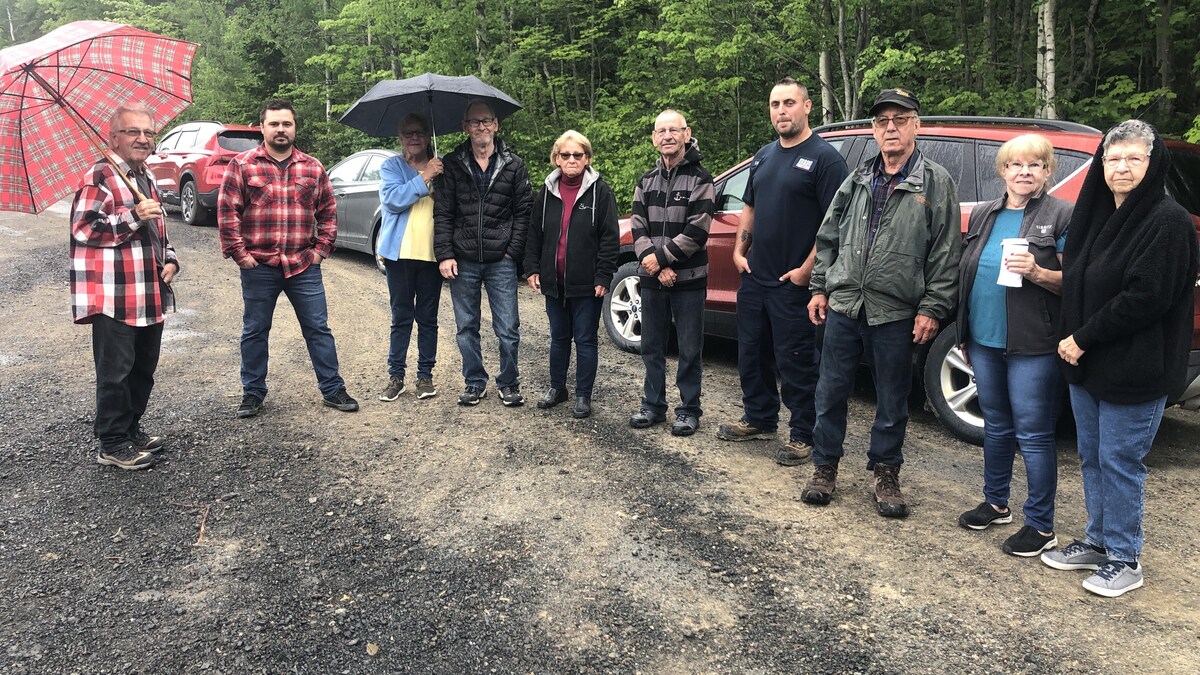 Un groupe d'une dizaine de personne, dont certains avec parapluie, devant des véhicules et des arbres.