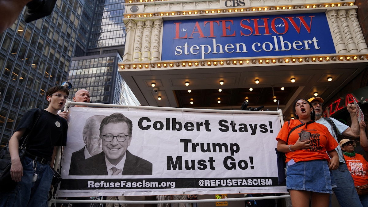 Devant la façade d'un immeuble illuminé, des manifestants brandissent une énorme banderole sur laquelle il est écrit en anglais : « Colbert doit rester! Trump doit partir! ».
