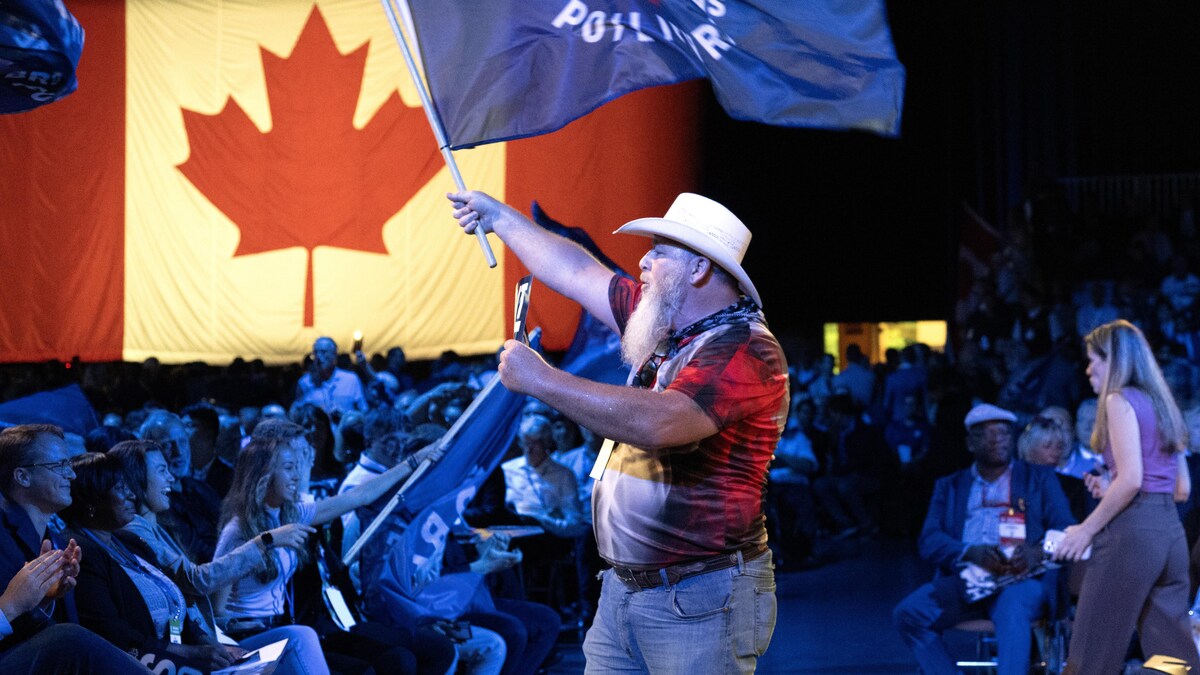 Un homme barbu coiffé d'un chapeau tient un drapeau en signe de soutien à Pierre Poilievre tandis qu'une assemblée de délégués conservateurs réagit à un discours.