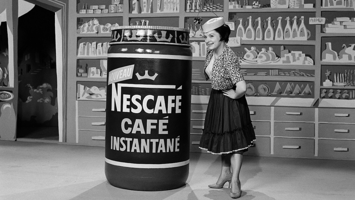 Mirielle Lachance dans un studio au décor d'épicerie à côté de contenant format géant de Nescafé.
