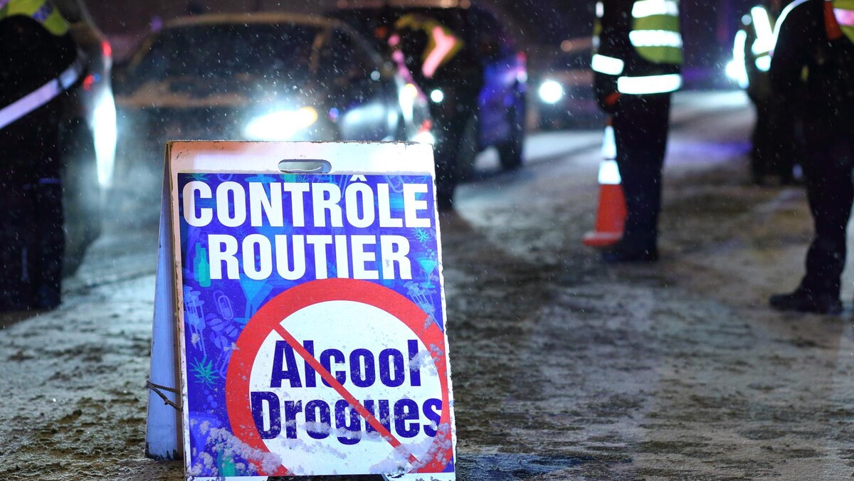 Des véhicules avancent lentement à un barrage routier, un soir d'hiver, sous la supervision de policiers.