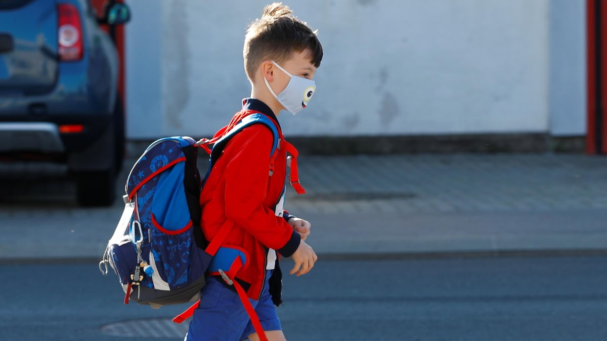 Un enfant en pantalon court porte un masque sur le visage. Il marche et a un sac à dos.