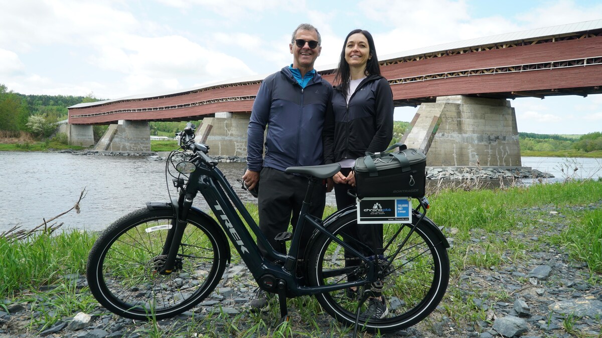 Un homme et une femme derrière un vélo électrique. À l'arrière-plan on peut apercevoir un long pont couvert rouge.