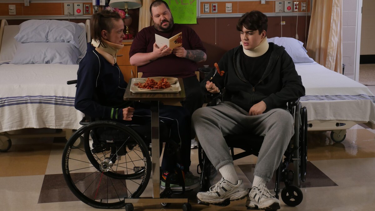 Deux personnes en fauteuil roulant mangent autour d’une table dans une chambre d’hôpital. Une troisième personne lit un livre.