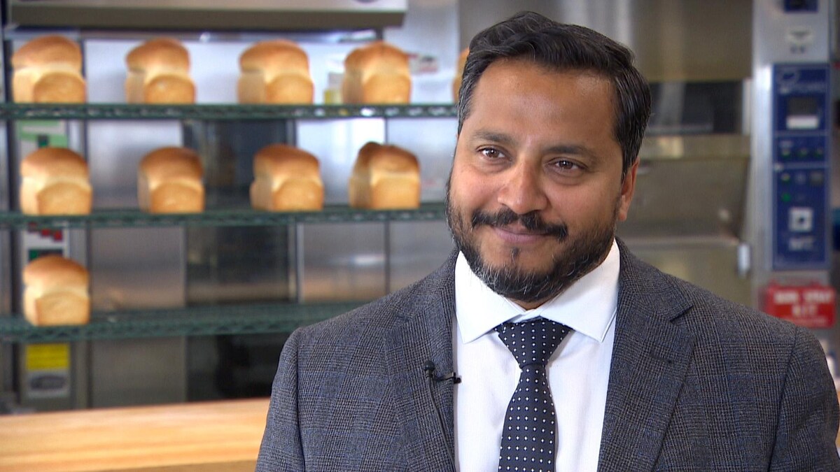 Un homme en complet cravate sourit dans les laboratoires de Cereals Canada. Derrière lui, un four en métal et une dizaine de miches de pain sur une étagère.