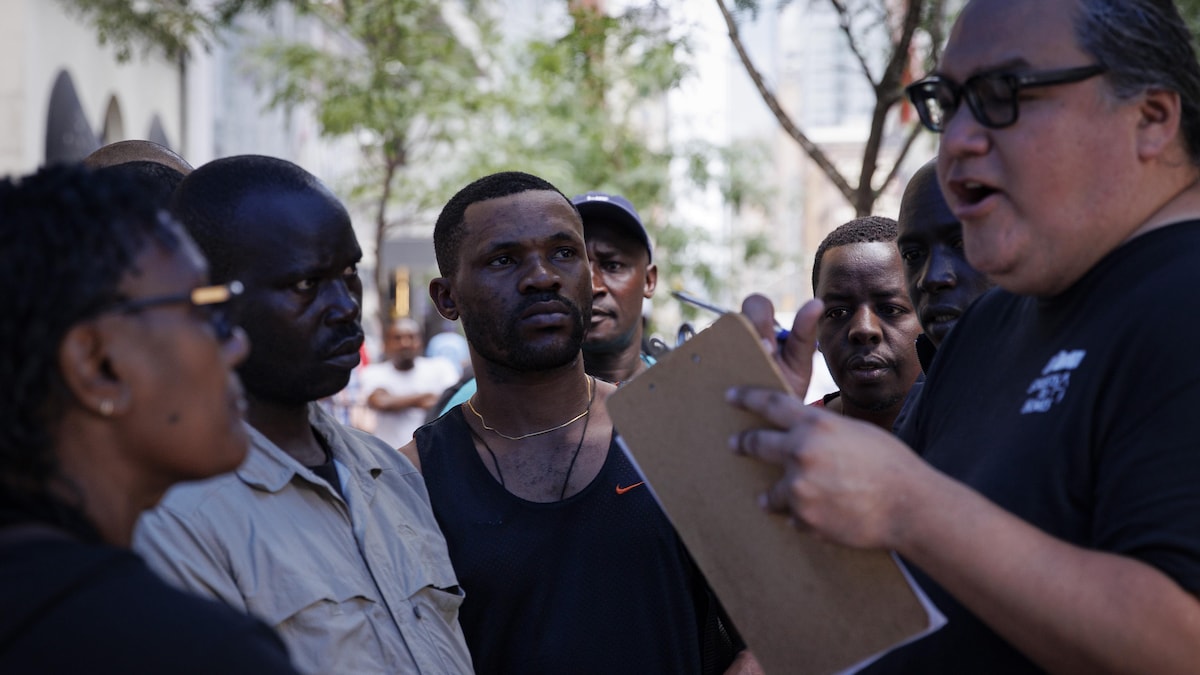 Un homme portant des lunettes tient un calepin et parle devant un groupe de demandeurs d'asile et de réfugiés.
