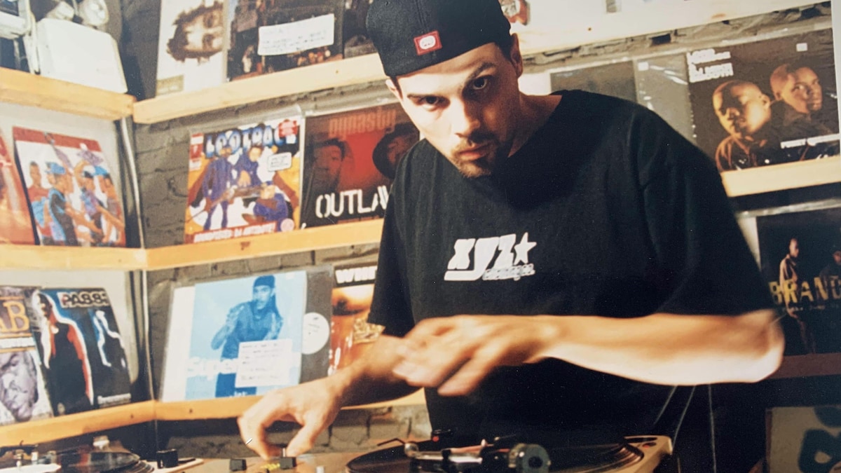 Un homme joue avec des platines de DJ devant une collection de vinyles exposés sur le mur. 