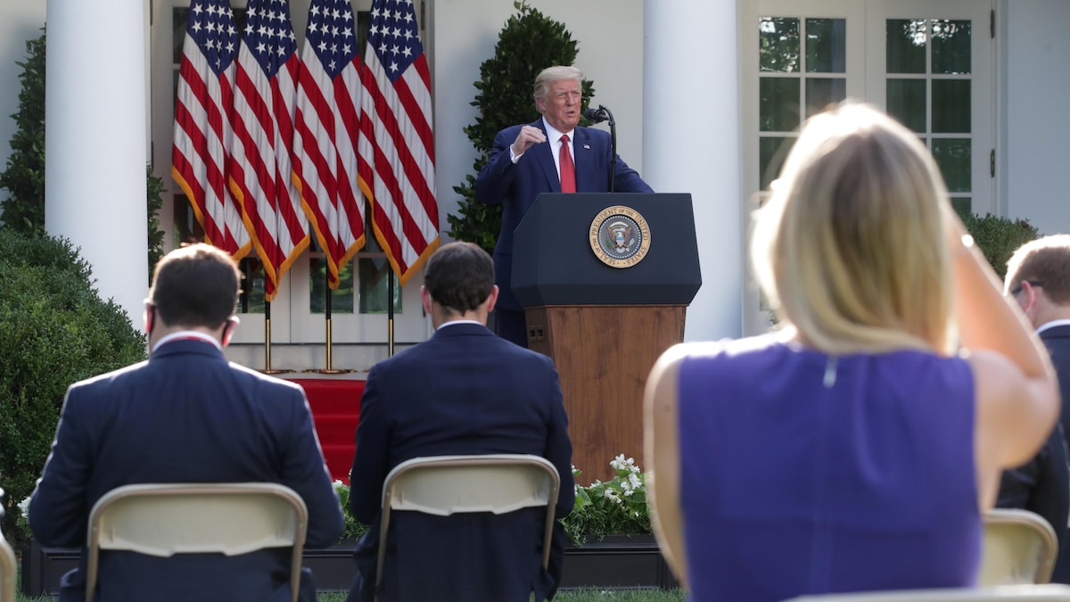 Devant des drapeaux américains, le président Trump, le bras levé, s'adresse aux journalistes, qu'on voit de dos.