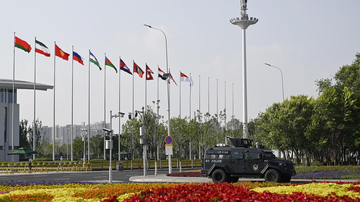 Un véhicule de la police chinoise se trouve devant des drapeaux des pays membres de l'OCS flottent au bout de leurs mâts devant un grand édifice.