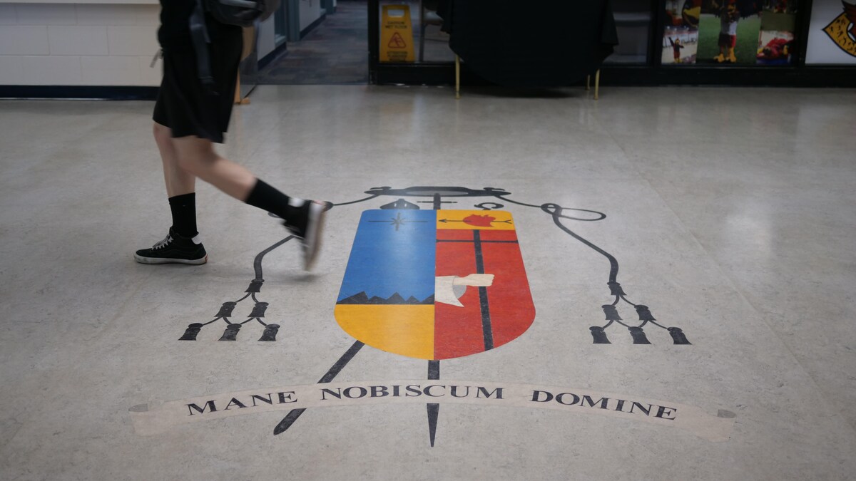 Un élève marche et au sol il y a les armoiries de l'école Bishop Carrol avec la locution latine Mane Nobiscum Domine (Reste avec nous Seigneur).