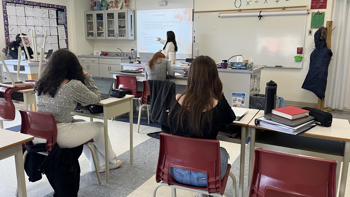Des élèves de dos assis à leurs pupitres écoutent une enseignante donner une leçon dans une salle d'école secondaire.