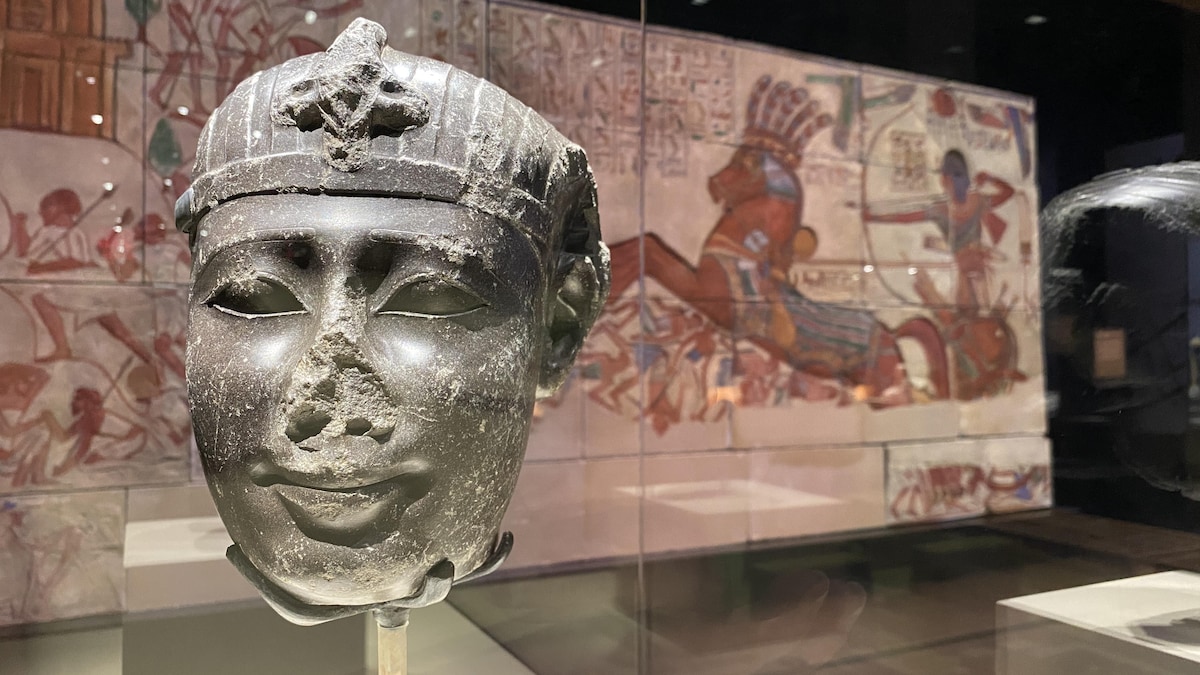 Une tête de pharaon en pierre est installée devant une fresque égyptienne. 