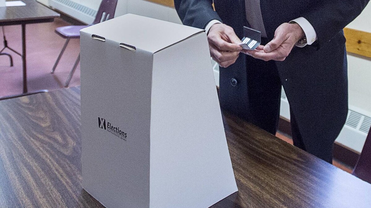Un homme debout devant une boîte de scrutin en carton tient un bulletin de vote.