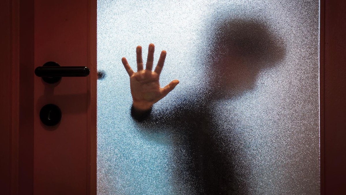 Un enfant derrière une fenêtre givrée dépose sa main contre la vitre et regarde au sol.