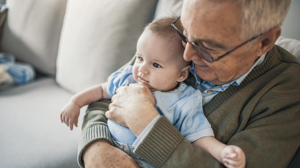 Un homme âgé, assis sur un sofa, sourit en tenant un bébé dans ses bras.