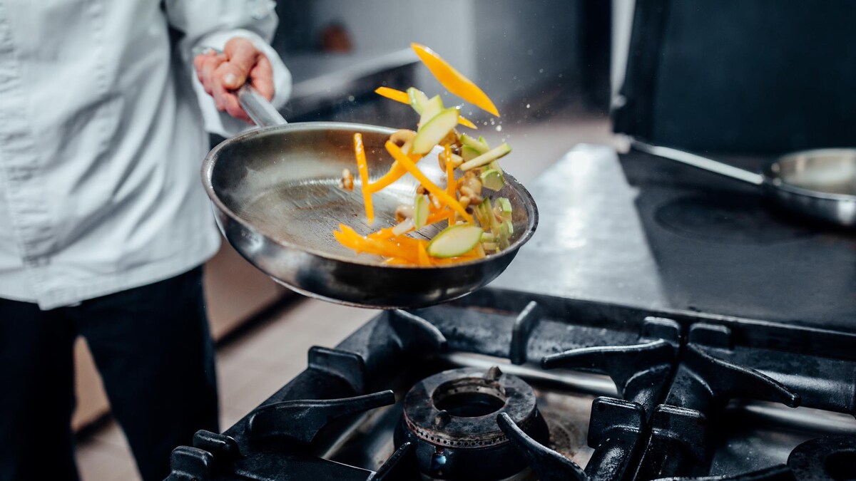 Une personne en habit de cuisinier fait sauter des légumes dans une poêle.