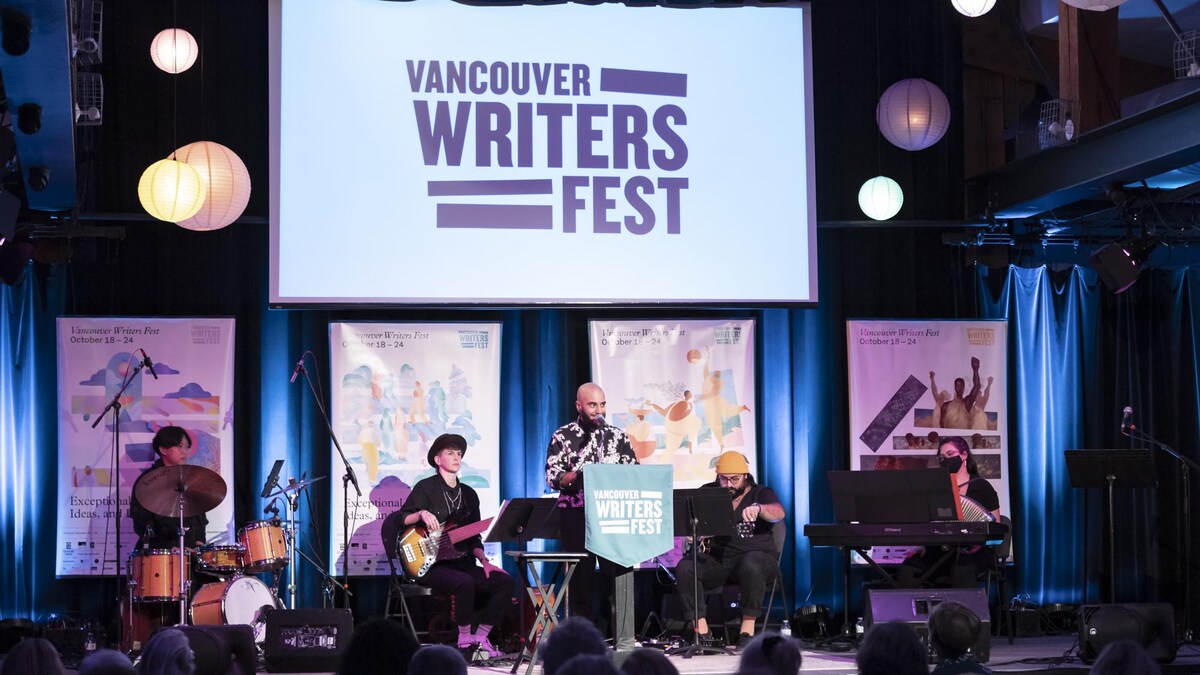 Une personne et un groupe de musique sur une scène devant un public. Au-dessus d'eux, il est écrit Vancouver Writers Fest.