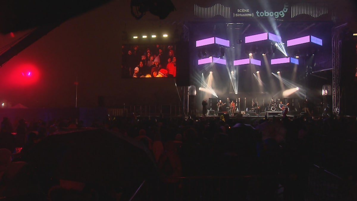 Un groupe de musique joue sur une scène extérieure, encadrée par des écrans géants et devant une foule de spectateurs garnie.