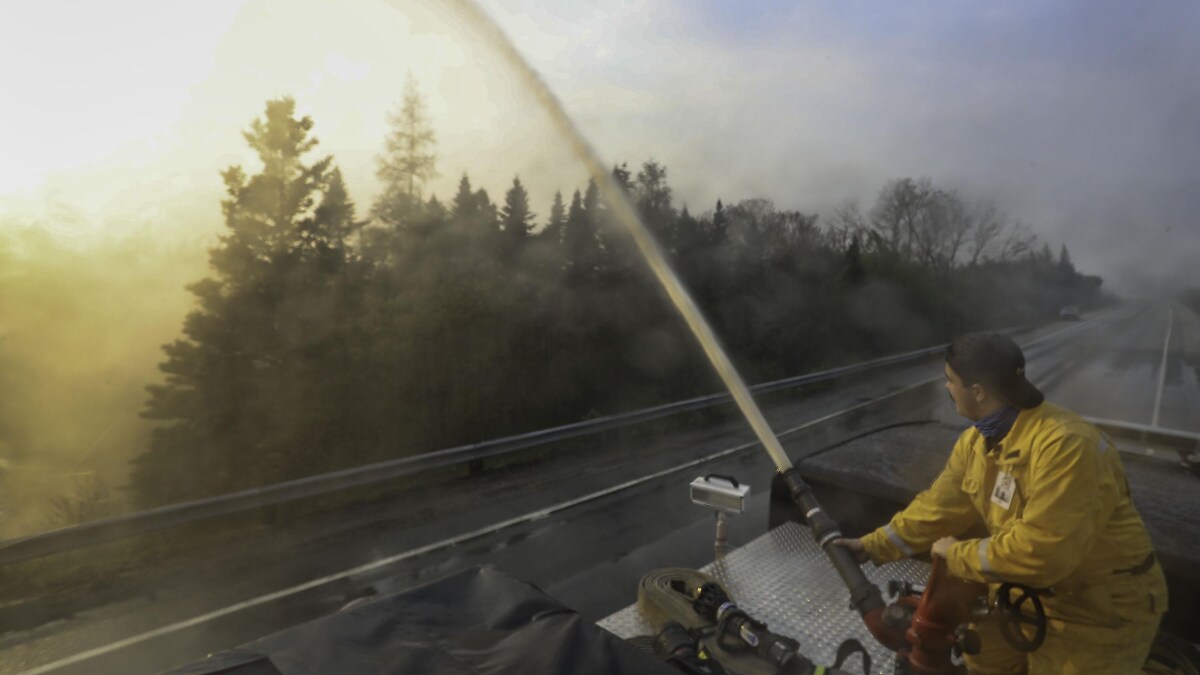 Un pompier vêtu d'un uniforme jaune arrose une zone boisée couverte de fumée, à partir d'un camion qui circule sur une autoroute.