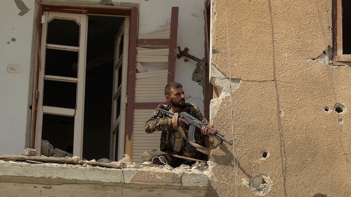Un combattant est accroupi sur le balcon d'un immeuble en ruines.