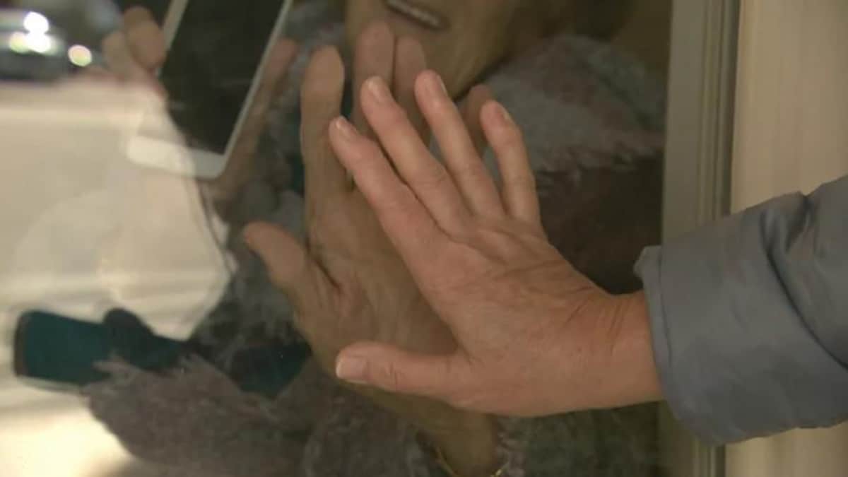 Une personne place sa main contre la main d'une autre personne qui se trouve dans un foyer de soins. Une vitre les sépare.