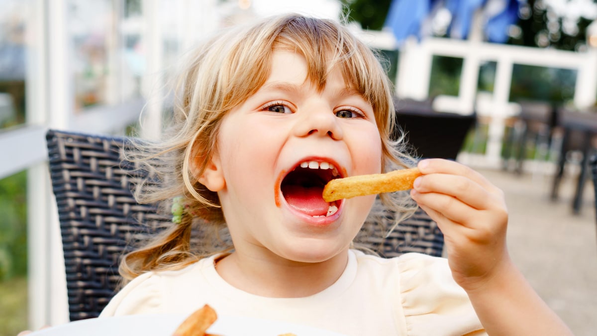 Une petite fille assise à l'extérieur devant une assiette de frites a la bouche grande ouverte et s'apprête à y faire entrer une frite.