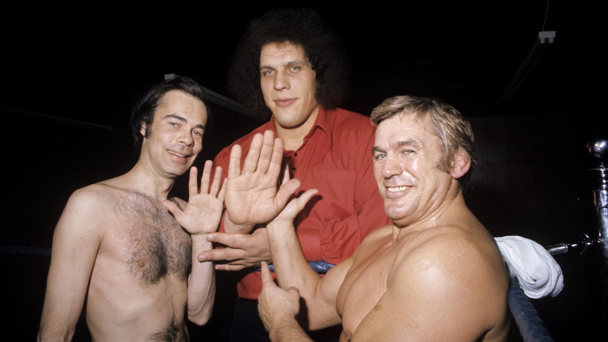 Sur un ring, le co-animateur Jacques Fauteux compare sa main à celle du
lutteur géant Ferré (André Roussimoff) sur laquelle pointe l'index de la main du
lutteur Édouard Carpentier.