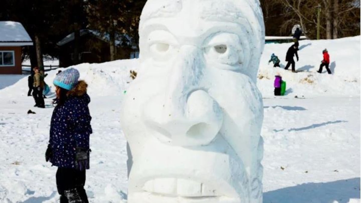 Une enfant se tient près d'une grande sculpture de neige qui représente un masque traditionnel autochtone.