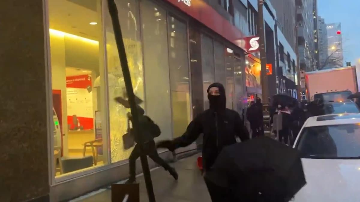 Un manifestant cagoulé brise la vitrine d'une succursale de la Banque Scotia sur la rue Sainte-Catherine, à Montréal, à l'aide d'une pancarte.
