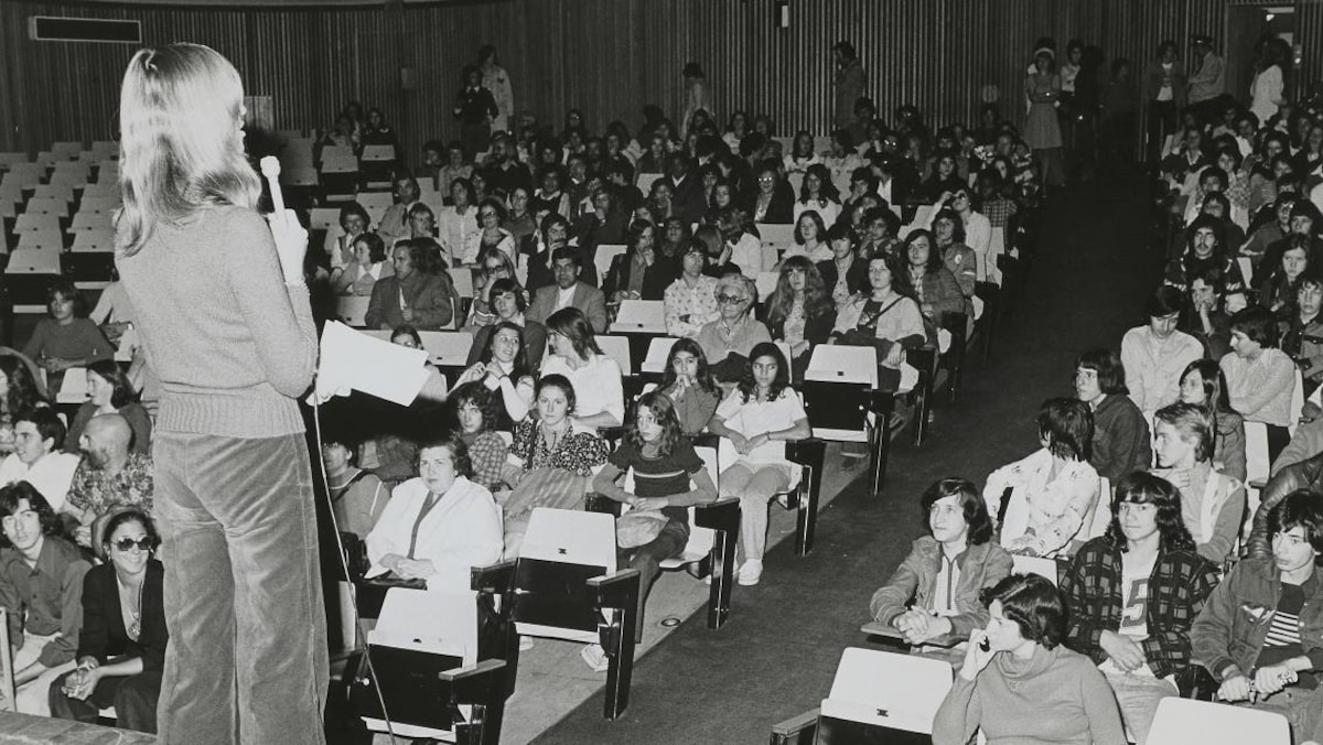 Une femme de dos tient un micro devant une foule assise dans une salle de spectacle.