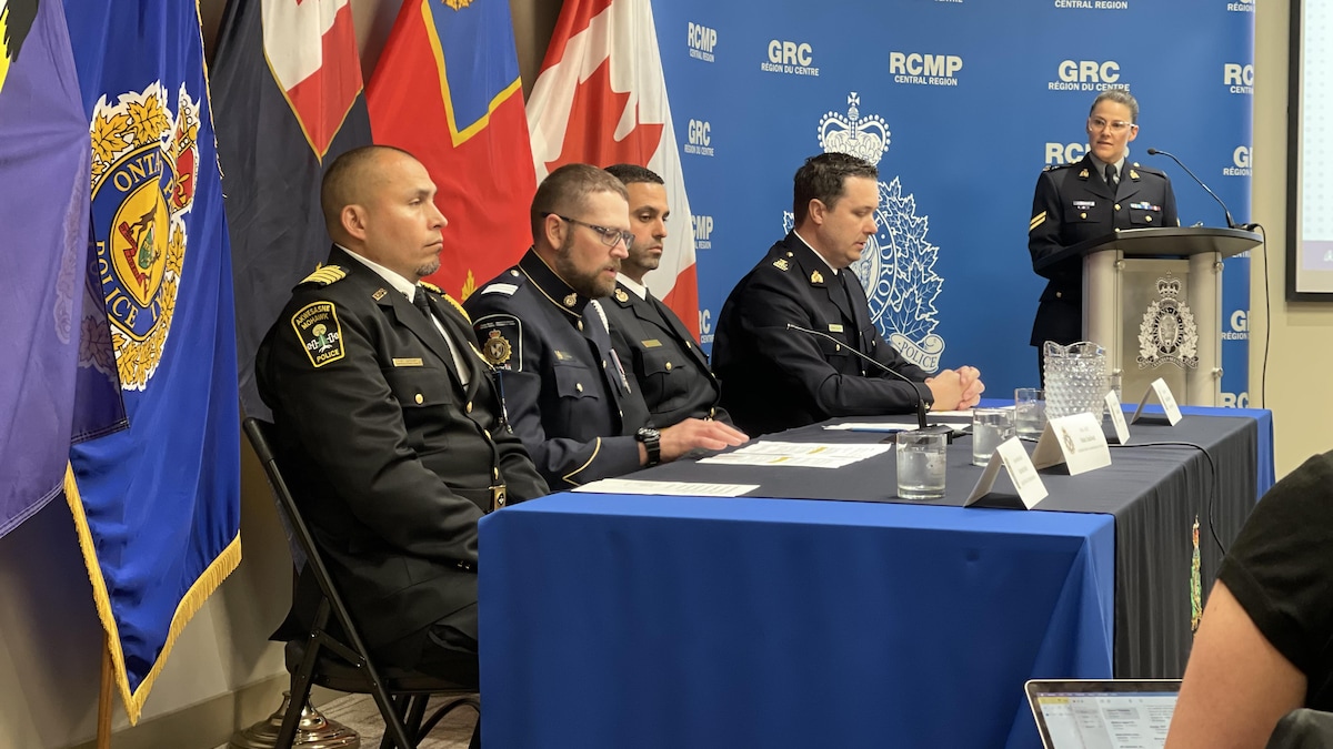 Quatre hommes en uniforme en conférence de presse assis à une table avec une femme, en uniforme, derrière un lutrin et un micro.