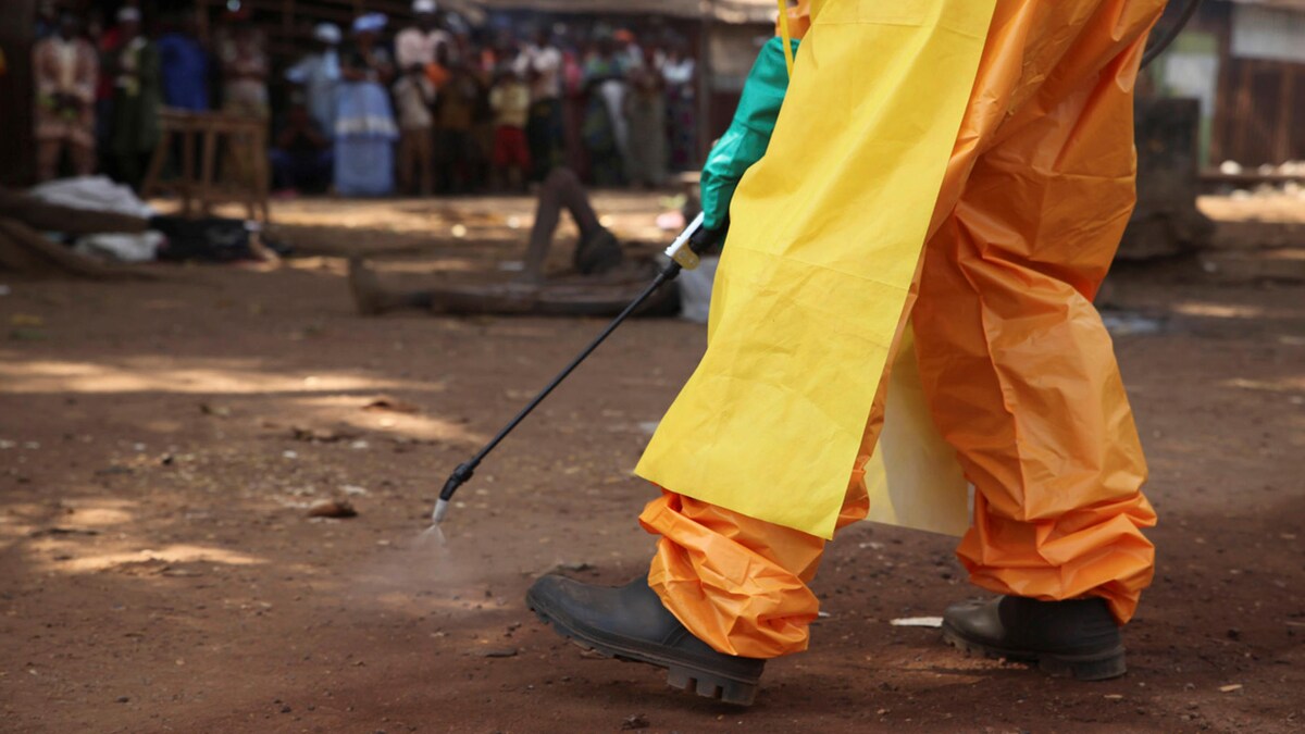 Une personne en habit de protection vaporise du désinfectant sur de la terre battue dans un village.