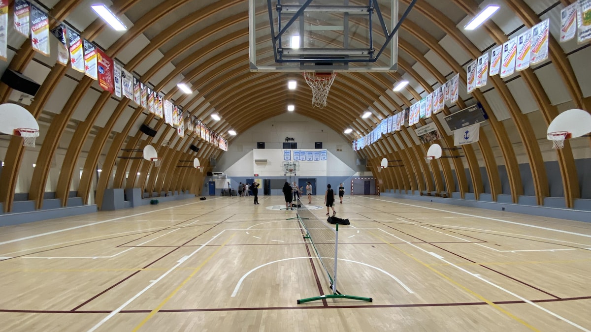 Photo intérieure d'un grand gymnase au plafond en arche, avec de multiples bannières de championnat accrochées dans les hauteurs et des paniers de basketball.