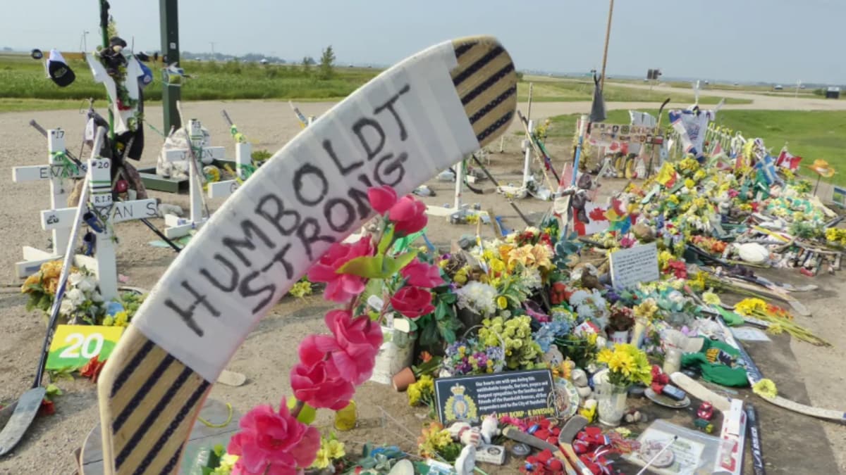 Un avant-plan sur un bâton de hockey avec les mots « Humboldt Strong » inscrits sur le ruban de la palette. Derrière, on voit des dizaines de fleurs de toutes les couleurs ainsi que d'autres objets pour commémorer les morts.